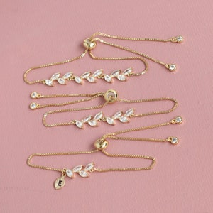 May include: Three gold-toned adjustable bracelets featuring a delicate leaf-shaped design set with clear, marquise-cut cubic zirconia stones. Each bracelet has a sliding bead closure and a thin box chain, with one bracelet including a small leaf charm with the letter E.