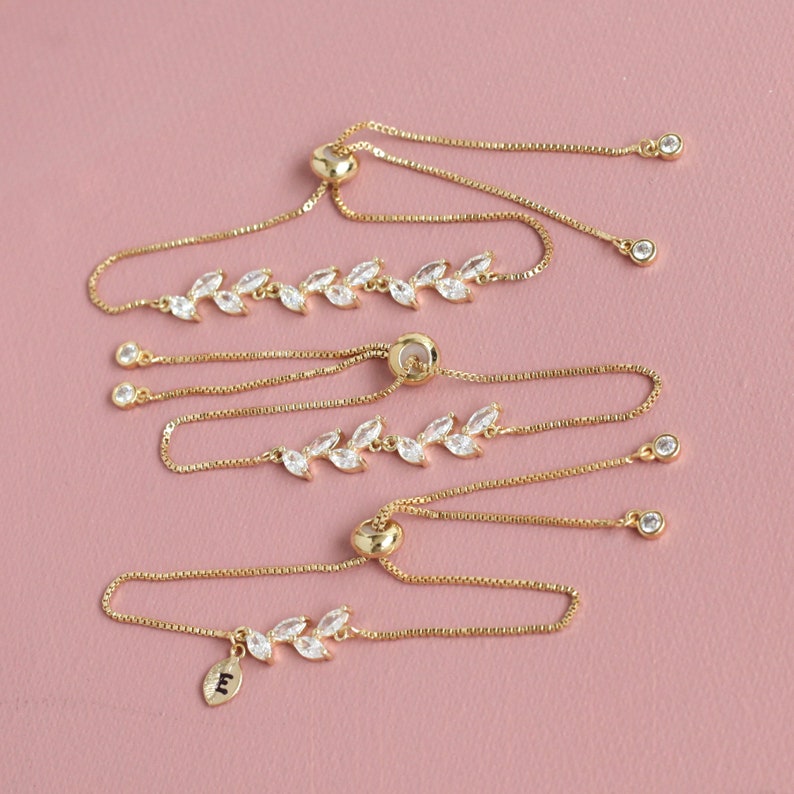 May include: Three gold-toned adjustable bracelets featuring a delicate leaf-shaped design set with clear, marquise-cut cubic zirconia stones. Each bracelet has a sliding bead closure and a thin box chain, with one bracelet including a small leaf charm with the letter E.