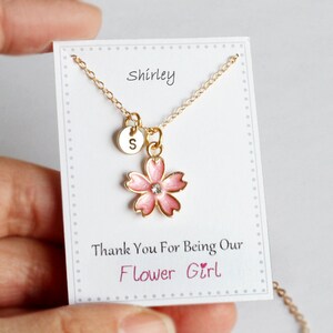 May include: A gold necklace with a pink flower charm and a small gold disc with the letter "S" on it. The necklace is on a white card with the text "Shirley" and "Thank You For Being Our Flower Girl".