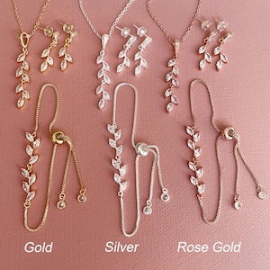 May include: Three jewelry sets in gold, silver, and rose gold tones, each featuring a necklace with a leaf-shaped pendant, matching drop earrings, and an adjustable bolo-style bracelet. Each piece is adorned with clear marquise-cut stones.