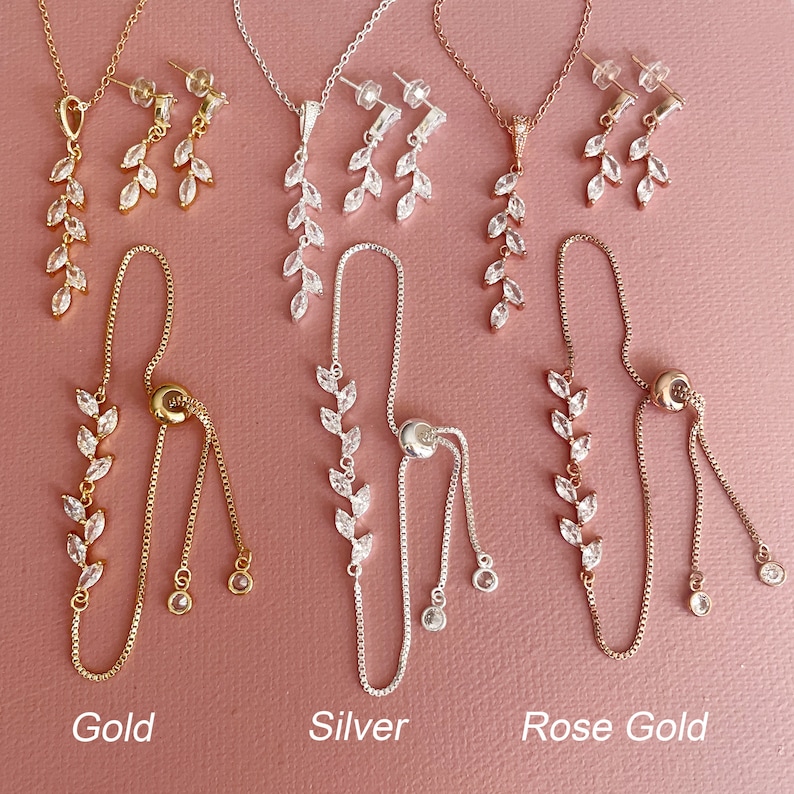 May include: Three jewelry sets in gold, silver, and rose gold tones, each featuring a necklace with a leaf-shaped pendant, matching drop earrings, and an adjustable bolo-style bracelet. Each piece is adorned with clear marquise-cut stones.