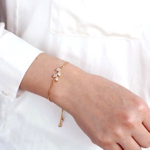K&ouml;nnte beinhalten: Ein goldfarbenes Kettenarmband mit einem zarten blattf&ouml;rmigen Design, das mit drei klaren Steinen im Marquise-Schliff verziert ist. Das Armband hat eine d&uuml;nne, verstellbare Kette mit zwei kleinen h&auml;ngenden Perlen am Ende.
