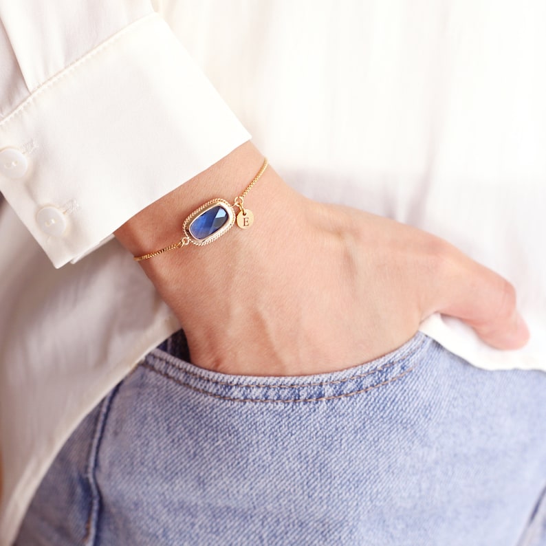 May include: A gold chain bracelet with a blue gemstone and a small charm with the letter "E" on it.