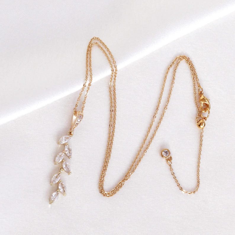 May include: A gold-toned necklace featuring a delicate chain and a vertical pendant composed of five marquise-cut clear stones arranged in a leaf-like pattern. The chain includes a small circular clasp and an adjustable length mechanism.