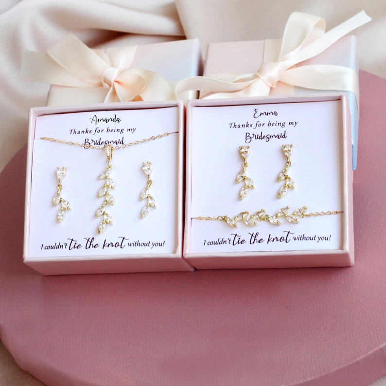 K&ouml;nnte beinhalten: Zwei hellrosa Geschenkboxen mit goldfarbenen Schmucksets aus blattf&ouml;rmigen, klaren Steinen. Die linke Box enth&auml;lt eine Halskette und Ohrringe, die rechte Ohrringe und ein Armband. Jede Karte tr&auml;gt den Text "Thanks for being my Bridesmaid" und "I couldn't tie the knot without you!" sowie die Namen Amanda und Emma.
