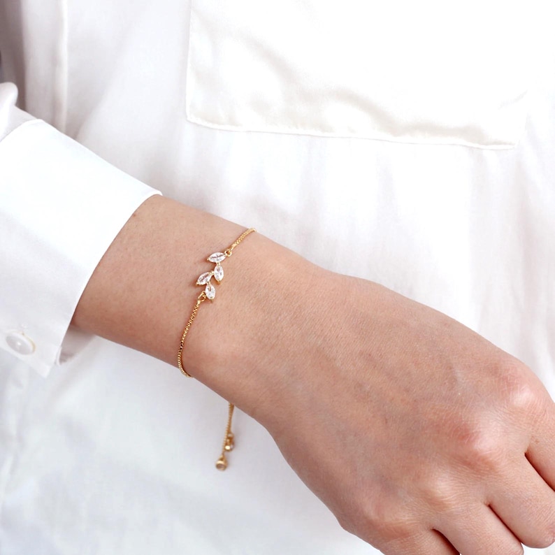 May include: A gold-toned chain bracelet featuring a delicate leaf-shaped design adorned with three clear marquise-cut stones. The bracelet includes a thin, adjustable chain mechanism with small dangling beads at the end for a secure fit.