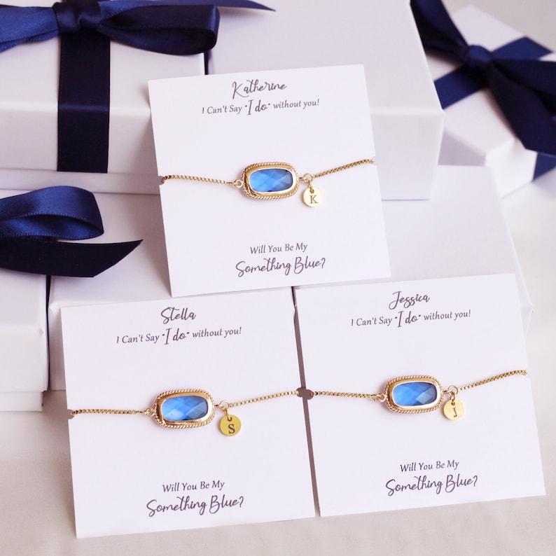 May include: Three bridesmaid gift boxes with blue and gold bracelets. Each box has a card with the text "Will You Be My Something Blue?" and a personalized message for each bridesmaid. The bracelets feature a blue gemstone and a gold initial charm.