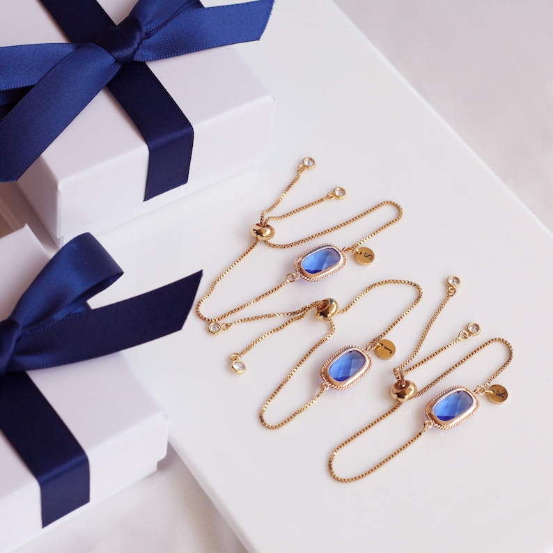 May include: Three gold chain bracelets with blue gemstone charms. Each bracelet has a small gold charm with a letter on it.