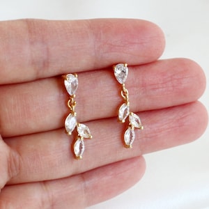 May include: A pair of gold-toned drop earrings featuring a branch-like design. Each earring consists of a single pear-shaped clear crystal at the top, connected by a small gold link to a cluster of three marquise-cut clear crystals arranged in a leaf pattern.
