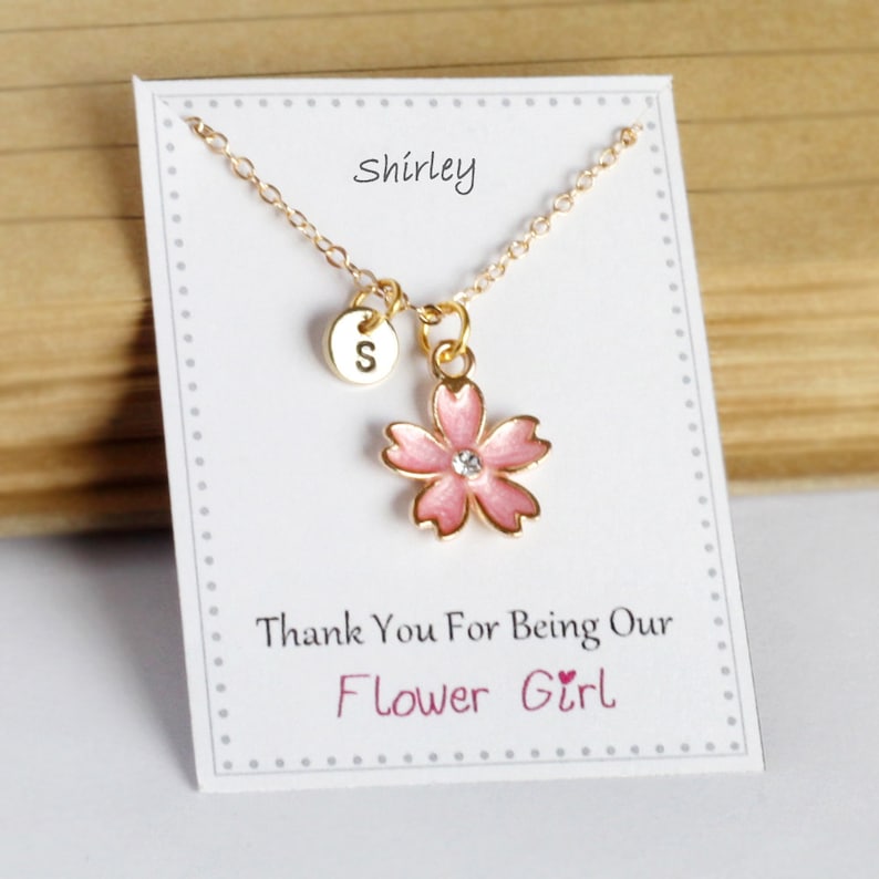 May include: Gold necklace with a pink flower charm and a small gold disc charm with the letter 'S' on it. The necklace is on a white card with the text 'Shirley' and 'Thank You For Being Our Flower Girl'.