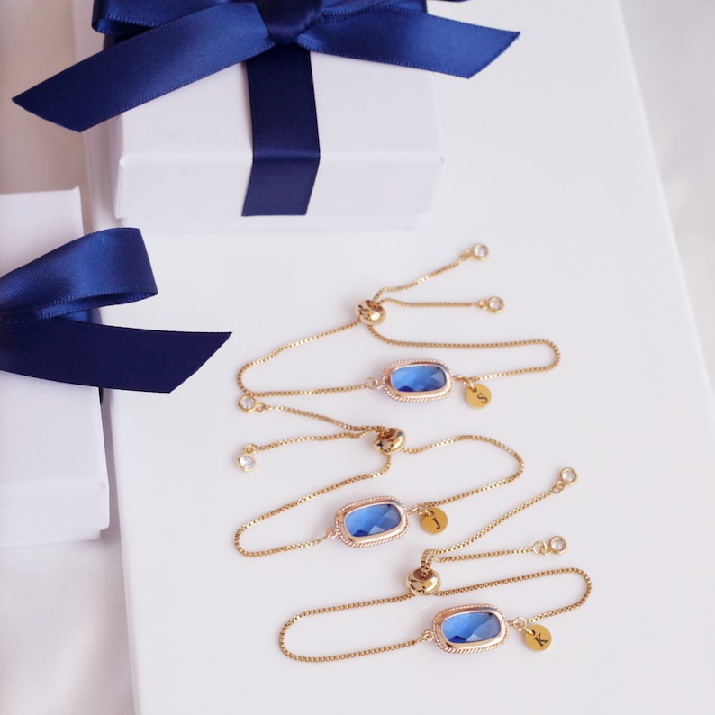 May include: Three gold chain bracelets with blue gemstone charms and initial charms. The charms are in the shape of a rectangle with rounded corners. The initials are "S", "J", and "K".