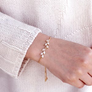 May include: A gold-toned bracelet featuring a delicate chain and a series of marquise-cut clear stones arranged in a leaf-like pattern. The bracelet includes a thin, adjustable chain segment that hangs down from the main decorative element.