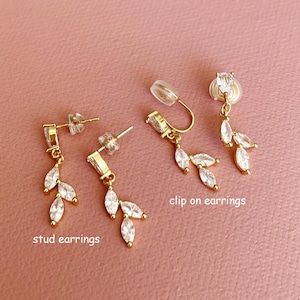 K&ouml;nnte beinhalten: Goldfarbene Ohrringe mit klaren, marquise-geschliffenen Steinen in blattartiger Anordnung. Das Set enth&auml;lt zwei Ohrstecker mit der Aufschrift stud earrings und zwei Clip-Ohrringe mit der Aufschrift clip on earrings, alle mit poliertem Metallfinish.