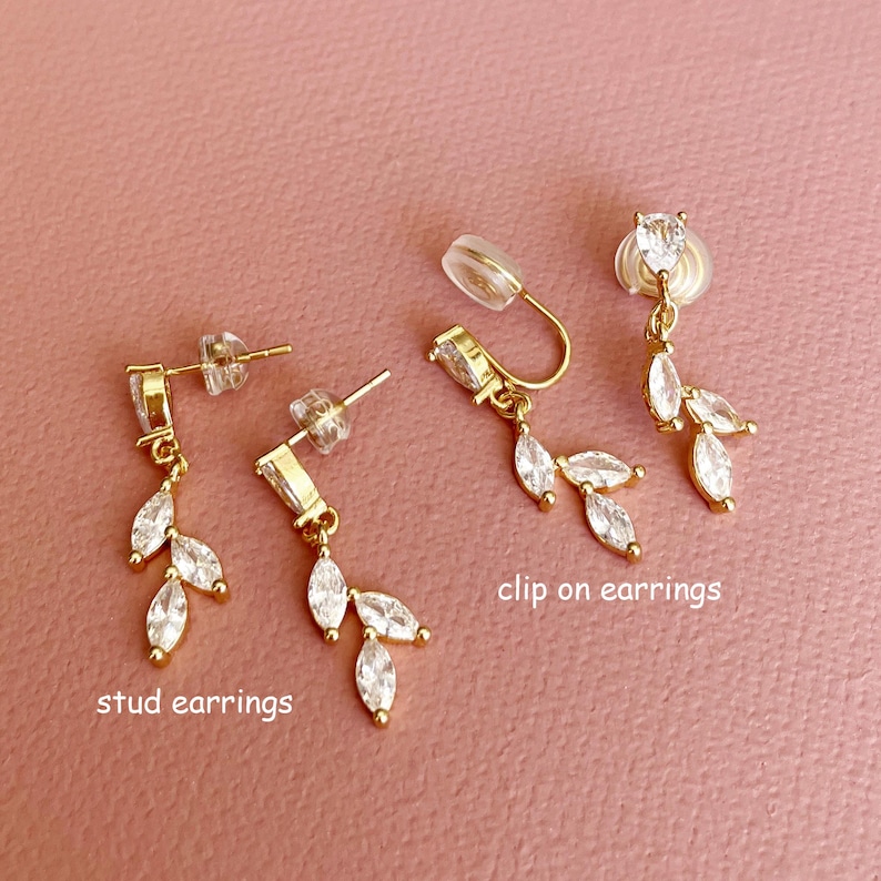 K&ouml;nnte beinhalten: Goldfarbene Ohrringe mit klaren, marquise-geschliffenen Steinen in blattartiger Anordnung. Das Set enth&auml;lt zwei Ohrstecker mit der Aufschrift stud earrings und zwei Clip-Ohrringe mit der Aufschrift clip on earrings, alle mit poliertem Metallfinish.