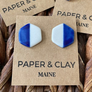 Könnte beinhalten: Ein Paar blaue und weiße Keramik-Hexagon-Ohrringe mit weißem Hintergrund und blauer Glasur auf der einen Hälfte jedes Ohrrings. Die Ohrringe werden auf einer braunen Karte mit dem Text "PAPER & CLAY MAINE" präsentiert.