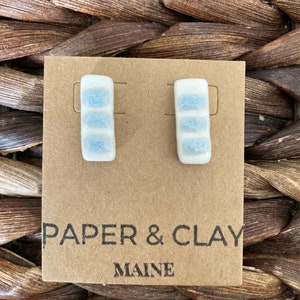 May include: A pair of small, rectangular ceramic earrings with a light blue and white speckled design. The earrings are displayed on a card that reads "PAPER & CLAY MAINE".