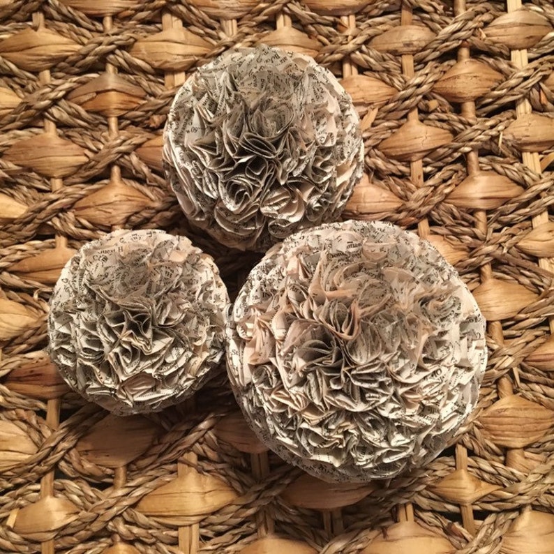 K&ouml;nnte beinhalten: Drei Papierblumenkugeln aus alten Buchseiten. Die Blumen sind wei&szlig; und cremefarben und auf einer geflochtenen Weidenoberfl&auml;che angeordnet.