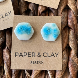 Könnte beinhalten: Ein Paar sechseckige Keramikohrringe mit weißer Basis und blauen Punkten. Die Ohrringe werden auf einer braunen Karte mit dem Text "PAPER & CLAY MAINE" präsentiert.