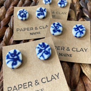 May include: A set of four pairs of blue and white ceramic earrings with a floral design. The earrings are displayed on brown cards with the text "Paper & Clay Maine".