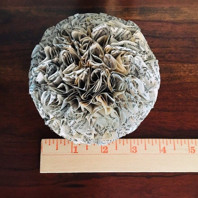 K&ouml;nnte beinhalten: Eine wei&szlig;e Papierblumenkugel aus Buchseiten. Die Blumenkugel hat einen Durchmesser von etwa 5 cm und liegt auf einer Holzoberfl&auml;che.