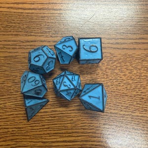 May include: A collection of seven blue polyhedral dice with black numbering and outlines. The dice are in various shapes, including a cube, a d20, and a d10. The dice are on a wooden surface.