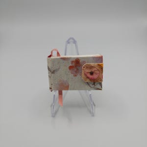 May include: A miniature book featuring a light-colored cover with a speckled pattern and orange floral motifs. The book is secured with a small, square tab closure decorated with a single flower and includes a thin, light pink ribbon bookmark.