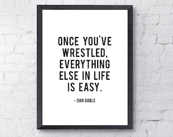 May include: Black and white motivational quote in a black frame on a white brick wall. The quote reads "Once you've wrestled, everything else in life is easy." - Dan Gable