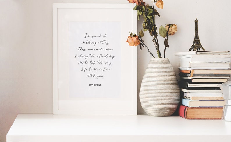 May include: A framed print with a white background and black text. The text reads: "I'm scared of walking out of this room and facing the rest of my whole life the way I feel when I'm with you." The text is signed "Dirty Dancing". The print is on a white shelf with a vase of dried flowers and a stack of books.