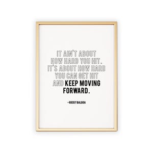 May include: A framed print with a gold-colored frame. The print features black text on a white background. The text reads: "IT AIN'T ABOUT HOW HARD YOU HIT. IT'S ABOUT HOW HARD YOU CAN GET HIT AND KEEP MOVING FORWARD." -ROCKY BALBOA.