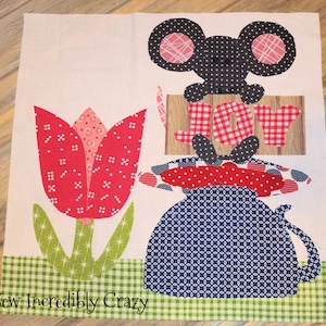 May include: A patchwork quilt block featuring a red and white tulip, a blue and white pitcher, a black mouse with pink ears, and a red and white checkered sign that says "JOY".