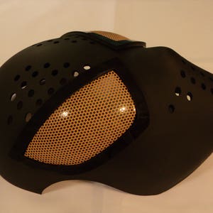 Puede incluir: M&aacute;scara de Halloween negra y naranja con un dise&ntilde;o de ojo de malla. La m&aacute;scara tiene una superficie texturizada y est&aacute; hecha de pl&aacute;stico duro.