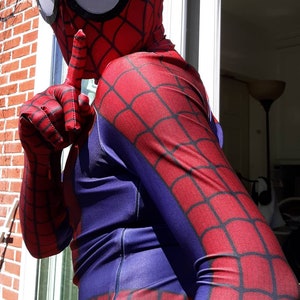 May include: A person wearing a red and blue Spiderman costume with a black spider web design. The person is holding their finger to their lips, as if to tell someone to be quiet.