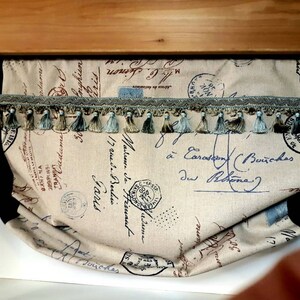 May include: A valance with a beige fabric background featuring a pattern of black and blue handwritten text and stamps. The valance has a black and beige trim with a blue and beige tassel fringe.