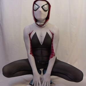 Puede incluir: Un traje de spandex blanco y rojo con una m&aacute;scara blanca y un dise&ntilde;o de ara&ntilde;a blanco y negro en los brazos. El traje lo lleva una persona en posici&oacute;n de cuclillas.