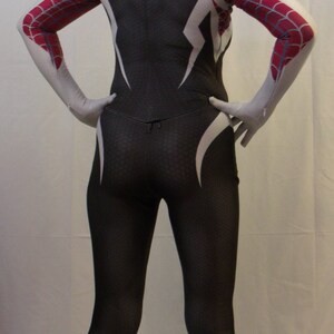 Puede incluir: Un traje de spandex blanco y negro con dise&ntilde;os de telara&ntilde;a roja y blanca. El traje tiene una capucha blanca con una m&aacute;scara blanca. La persona que lleva el traje est&aacute; de pie sobre una superficie blanca.