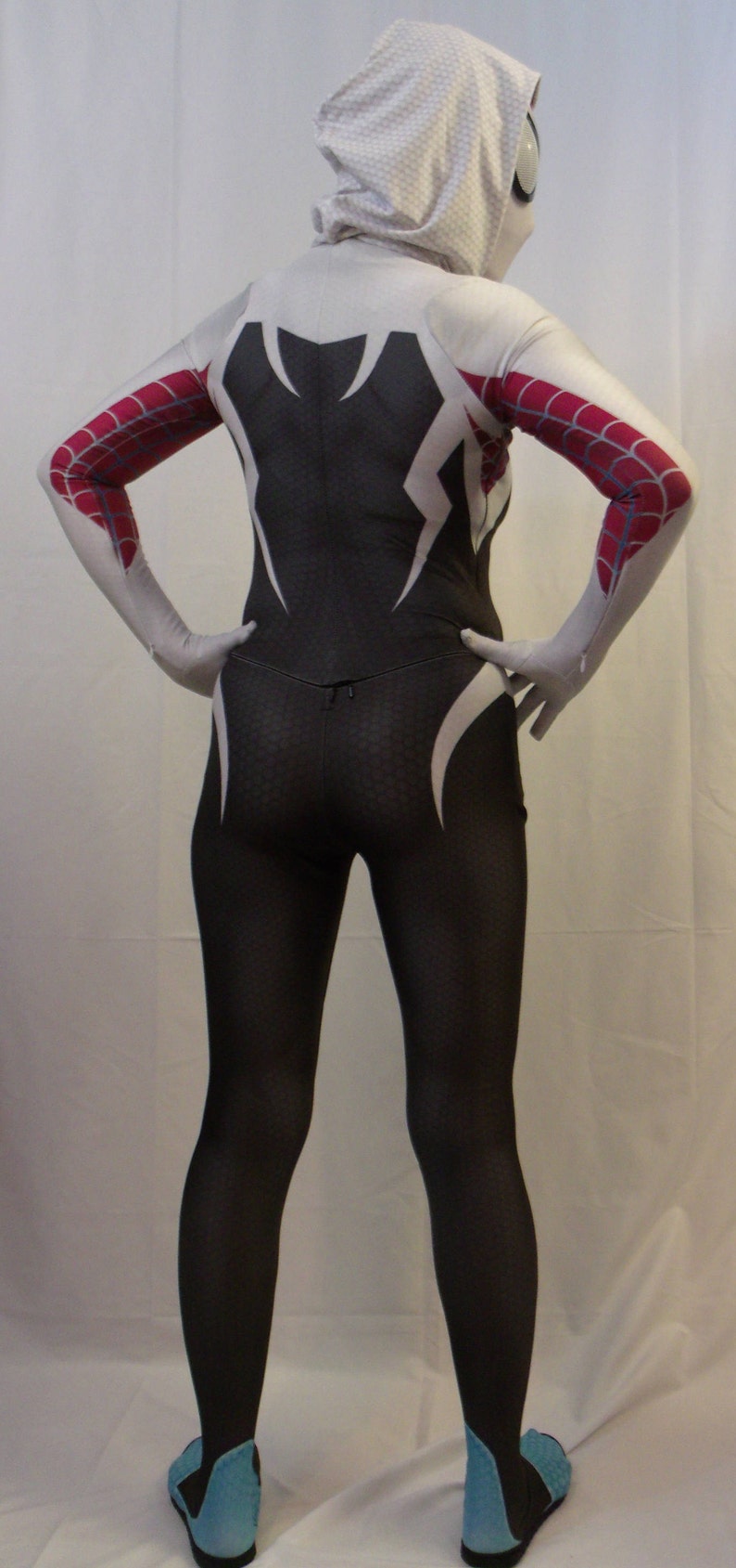 Puede incluir: Un traje de spandex blanco y negro con dise&ntilde;os de telara&ntilde;a roja y blanca. El traje tiene una capucha blanca con una m&aacute;scara blanca. La persona que lleva el traje est&aacute; de pie sobre una superficie blanca.