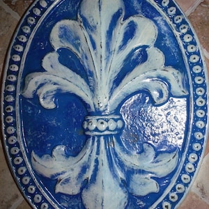 May include: A blue and white ceramic wall plaque with a fleur-de-lis design. The plaque is oval shaped and has a beaded border.