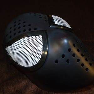 Może przedstawiać: Czarna maska ​​z tworzywa sztucznego z białym wzorem siatkowym. Maska ​​ma szereg małych otwor&oacute;w wok&oacute;ł krawędzi.