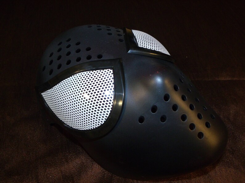 Może przedstawiać: Czarna maska ​​z tworzywa sztucznego z białym wzorem siatkowym. Maska ​​ma szereg małych otwor&oacute;w wok&oacute;ł krawędzi.