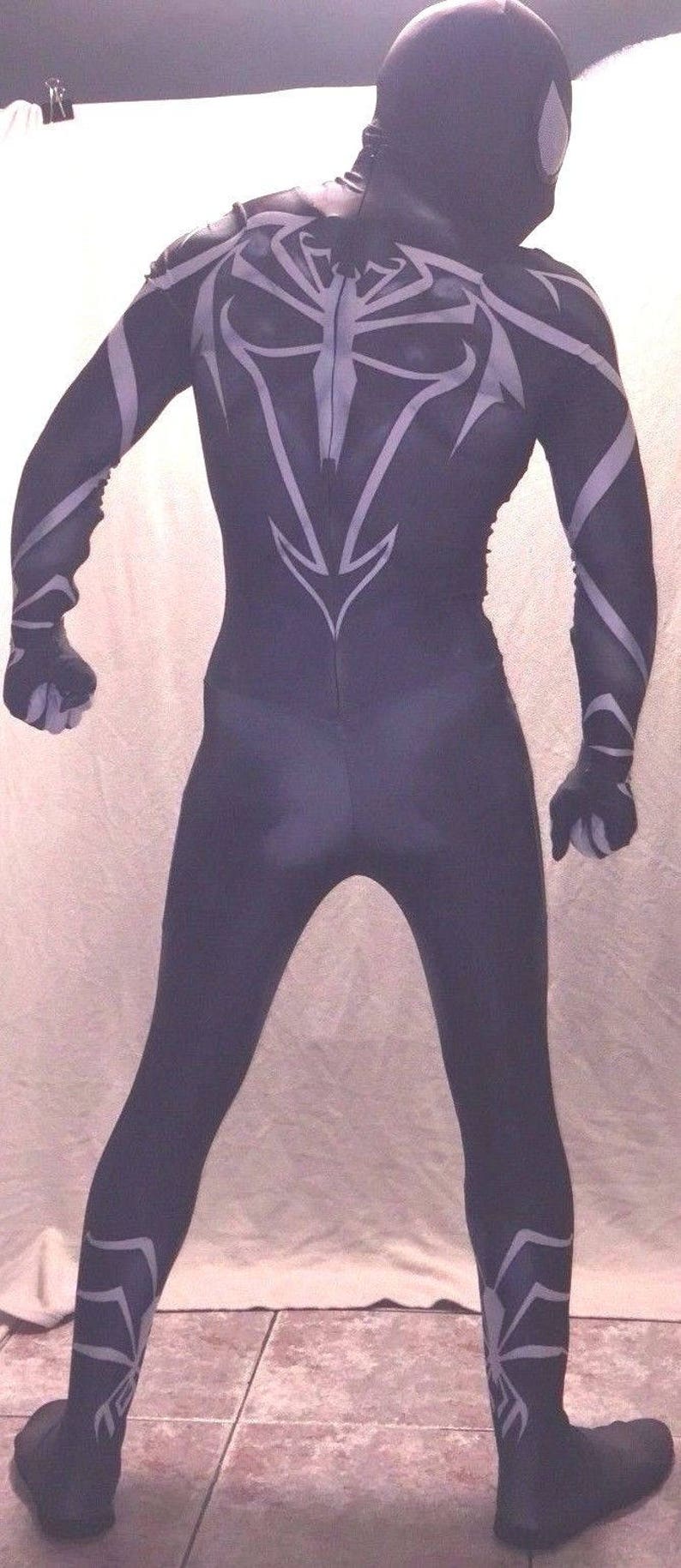 Może przedstawiać: Czarno-biały kombinezon z elastycznego materiału z wzorem pająka na plecach i nogach. Kombinezon ma czarną maskę z białym wzorem pająka.