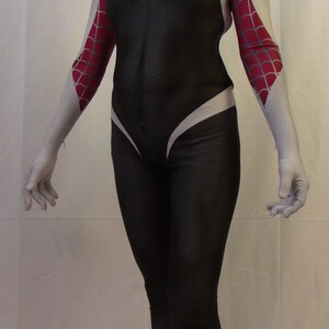 Puede incluir: Un disfraz de ara&ntilde;a blanco y rojo con una m&aacute;scara blanca y un mono negro. El disfraz tiene un s&iacute;mbolo de ara&ntilde;a blanco en el pecho y una telara&ntilde;a de ara&ntilde;a roja en los brazos. La persona que lleva el disfraz est&aacute; de pie sobre un fondo blanco.