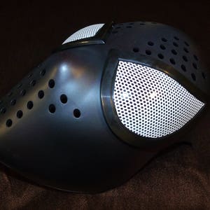 May include: A black plastic mask with a white mesh eye covering. The mask has a textured surface and multiple small holes.