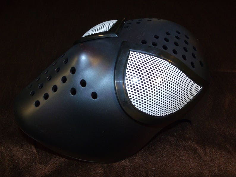 May include: A black plastic mask with a white mesh eye covering. The mask has a textured surface and multiple small holes.