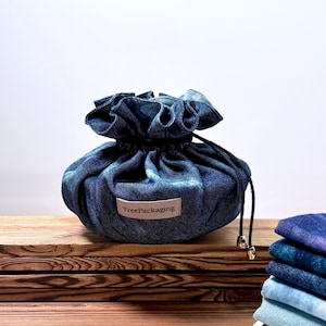 Puede incluir: Una bolsa de mezclilla con cordón y parte superior con volantes, sobre una superficie de madera. La bolsa tiene una pequeña etiqueta que dice "TreePackaging". Se ve una pila de tela de mezclilla doblada. La bolsa mide unos 15 cm de diámetro.