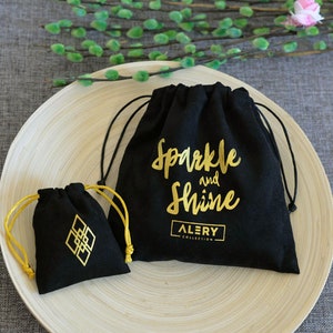 May include: Two black velvet drawstring bags with gold lettering. The larger bag says "Sparkle and Shine" with the word "ALERY" below. The smaller bag has a geometric design.