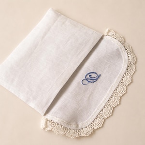 May include: A white linen handkerchief featuring a rounded edge trimmed with intricate cream-colored lace. The fabric is folded, revealing a blue embroidered cursive letter S on the surface. The textile has a visible weave and a soft texture.