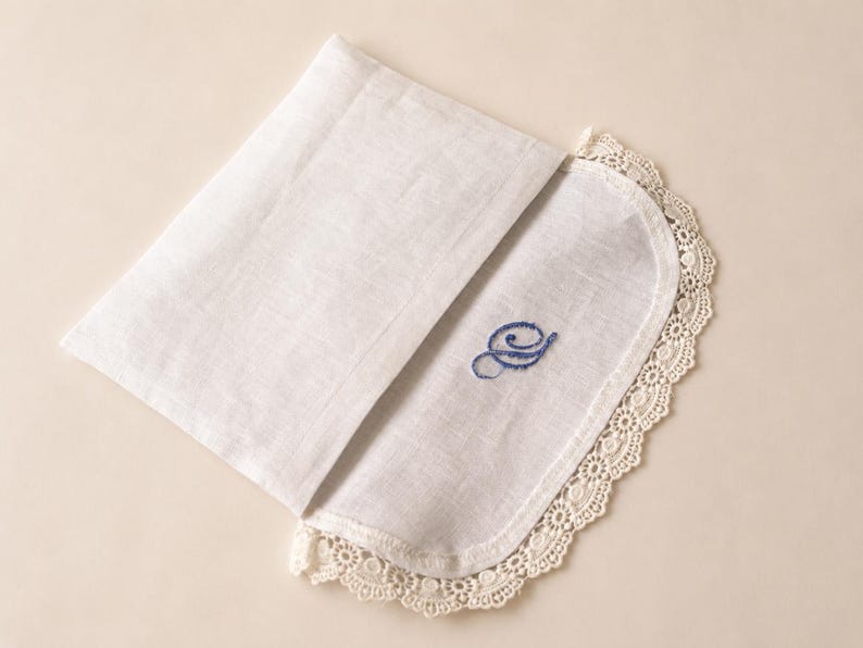 May include: A white linen handkerchief featuring a rounded edge trimmed with intricate cream-colored lace. The fabric is folded, revealing a blue embroidered cursive letter S on the surface. The textile has a visible weave and a soft texture.