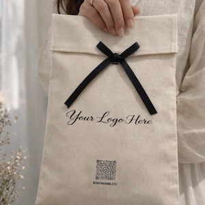 May include: A natural canvas bag with a black ribbon closure. The bag features the text "Your Logo Here" in a script font, along with a QR code and Instagram handle. The bag is held up by a person.