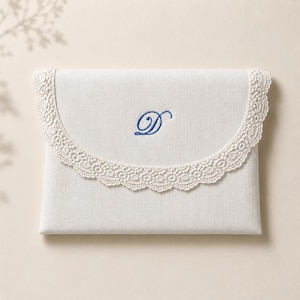 May include: A rectangular off-white fabric envelope pouch featuring a curved flap edge trimmed with intricate cream-colored lace. The center of the flap displays a single embroidered letter D in a deep blue cursive script font.