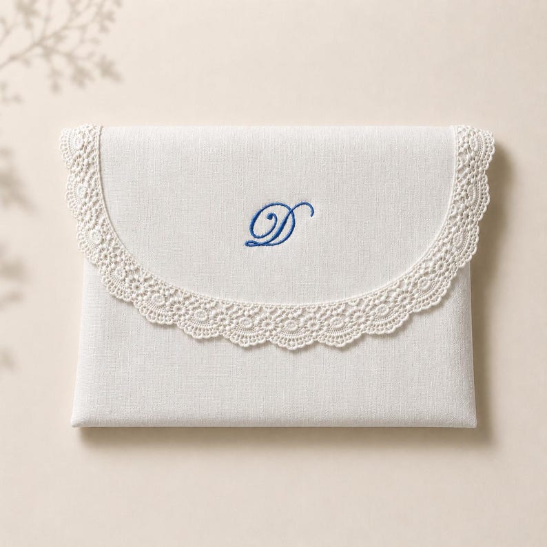 May include: A rectangular off-white fabric envelope pouch featuring a curved flap edge trimmed with intricate cream-colored lace. The center of the flap displays a single embroidered letter D in a deep blue cursive script font.
